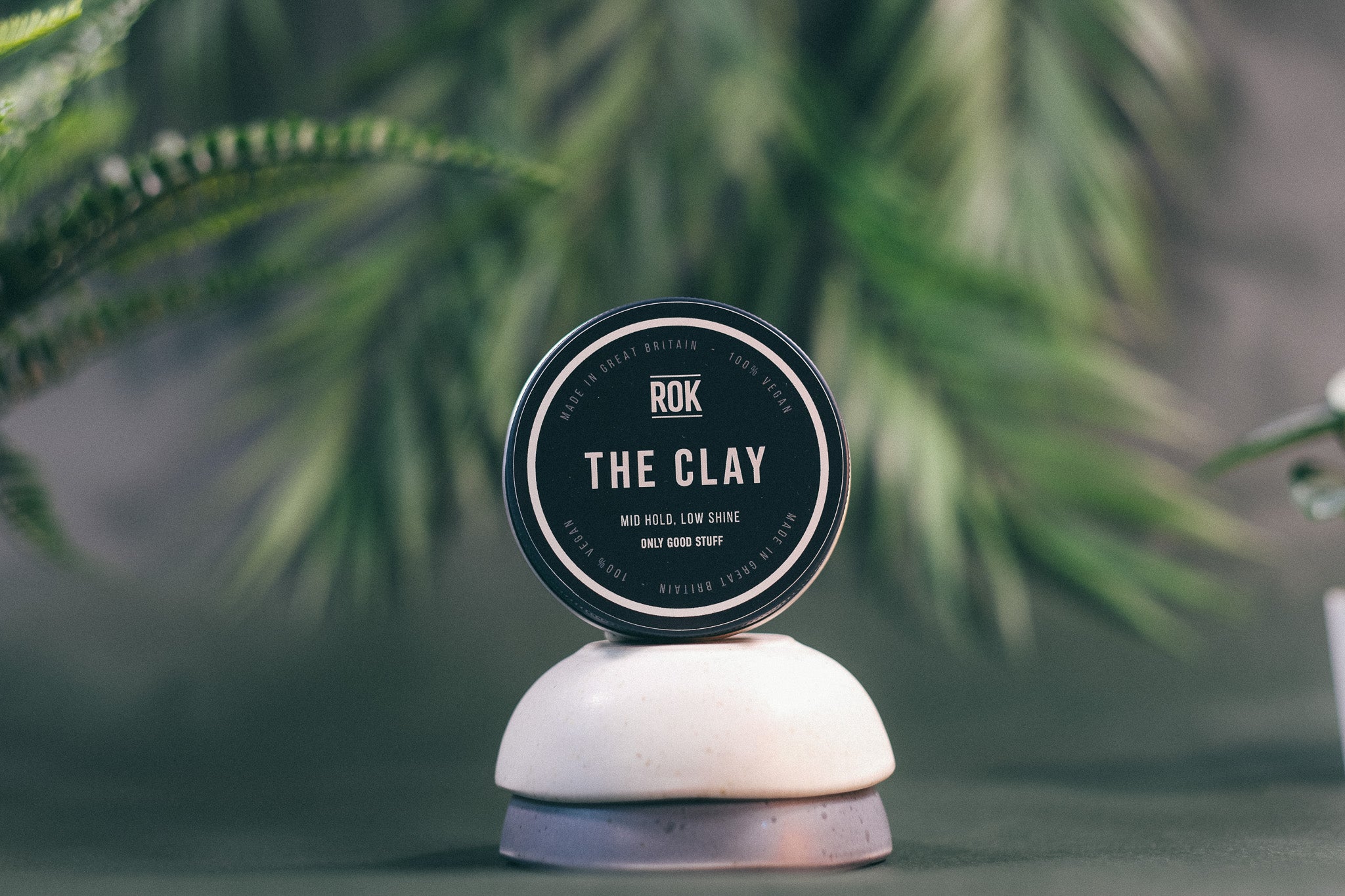 ROK The Clay tin on white stand with blurred green palm leaves background – vegan hair clay UK