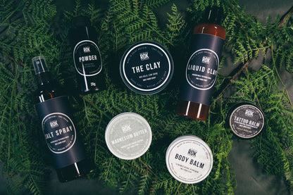 Full ROK Products range on green fern background including The Clay, Liquid Clay, Powder, Salt Spray, Body Balm and Tattoo Balm