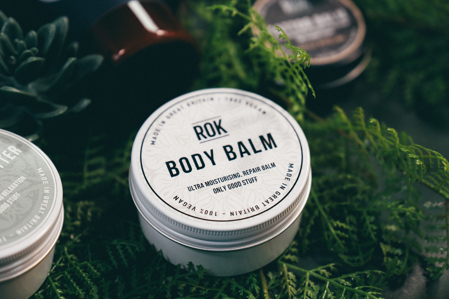 ROK Body Balm tin surrounded by green fern foliage with other ROK products – vegan body balm UK