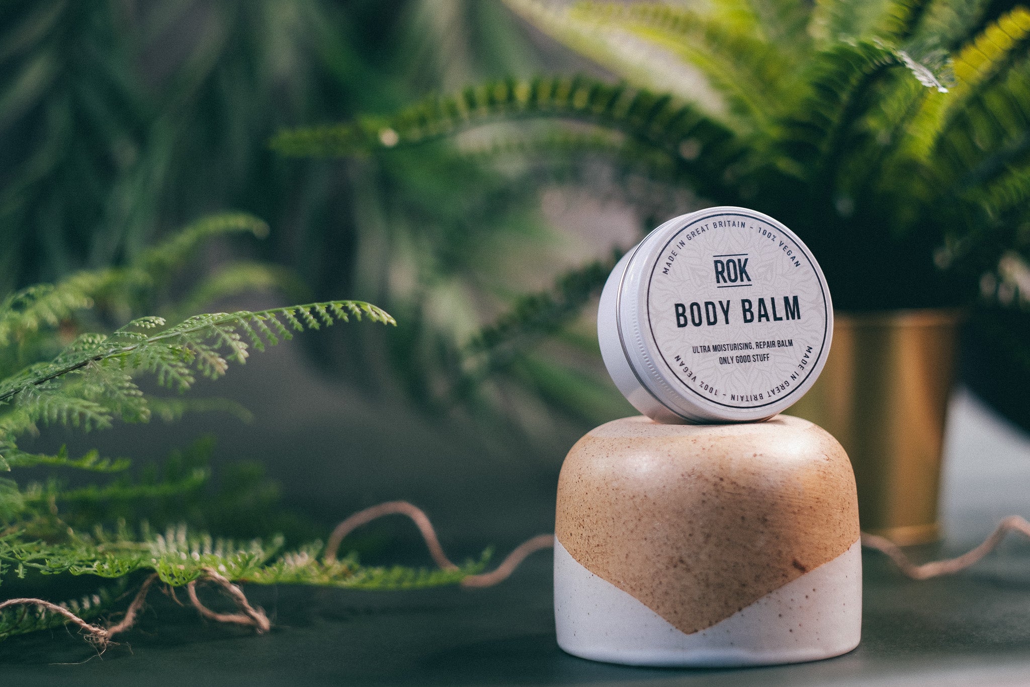 ROK Body Balm tin on stone pedestal with fern and brass pot background – vegan moisturising balm UK