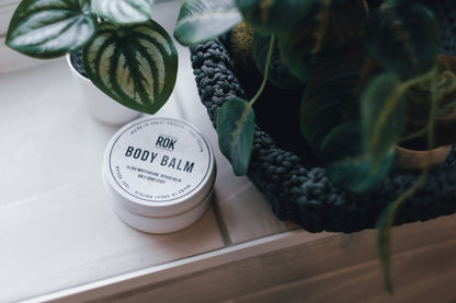 ROK Body Balm tin on wooden shelf with woven basket and tropical plants – vegan body moisturiser UK