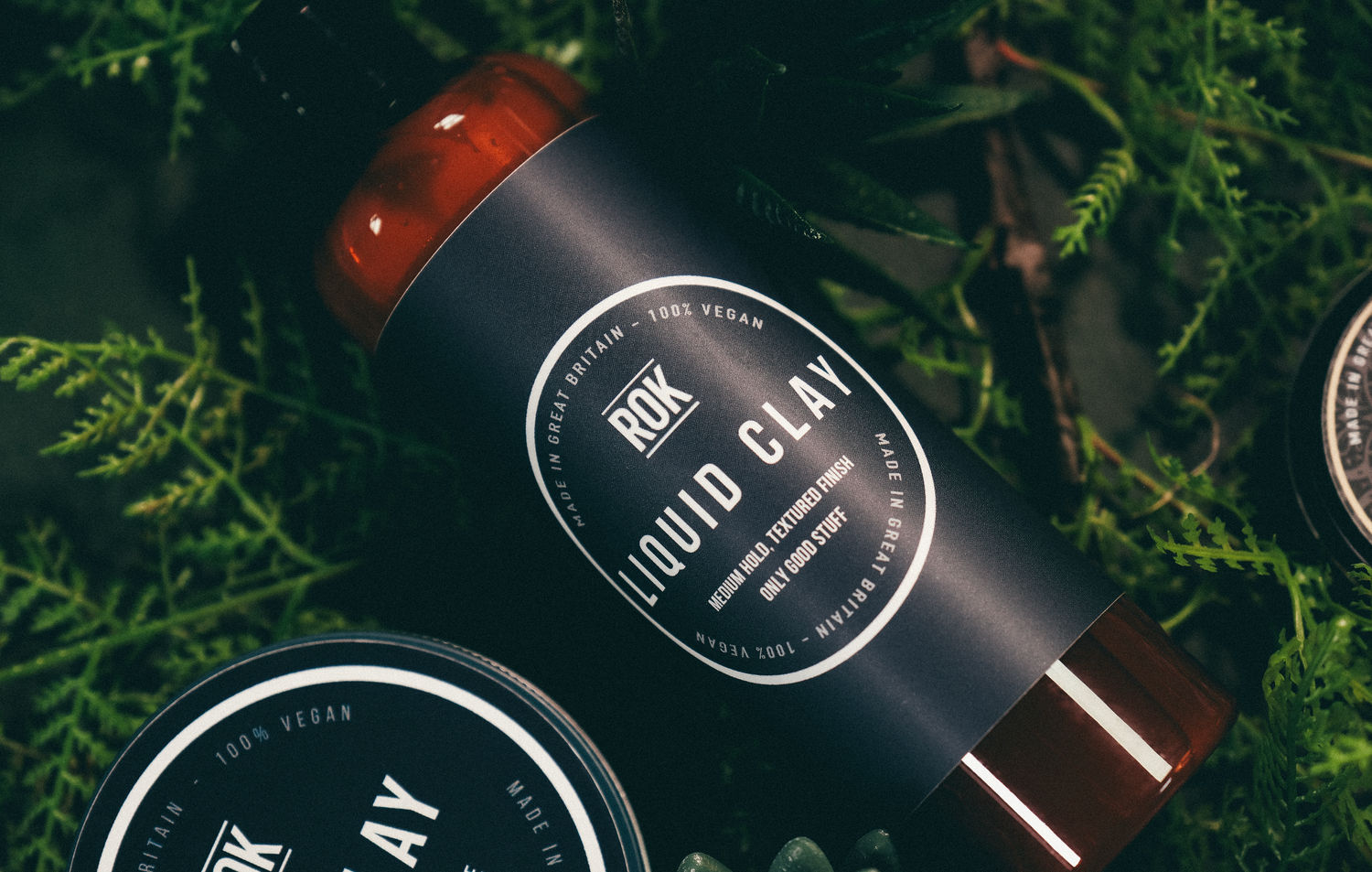 ROK Liquid Clay bottle and The Clay tin on green fern foliage – vegan hair styling products UK